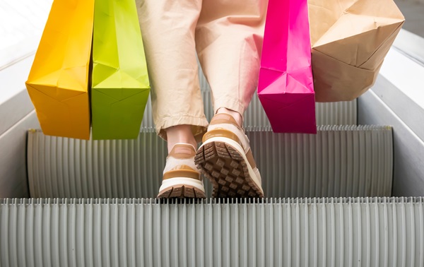The lower half of a person descending an escalator, wearing light pants and brown sneakers, holds bright pink, yellow, green, and brown shopping bags hanging down to the moving metal steps.