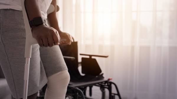 A person with a bandaged leg using a crutch to stand in a room with a wheelchair and a sunlit window.