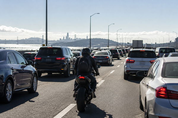 A motorcyclist lane splits between stopped vehicles in heavy freeway traffic with the San Francisco Bay Bridge and city skyline visible in the background, depicting the risks motorcyclists face on California's congested and dangerous roadways.