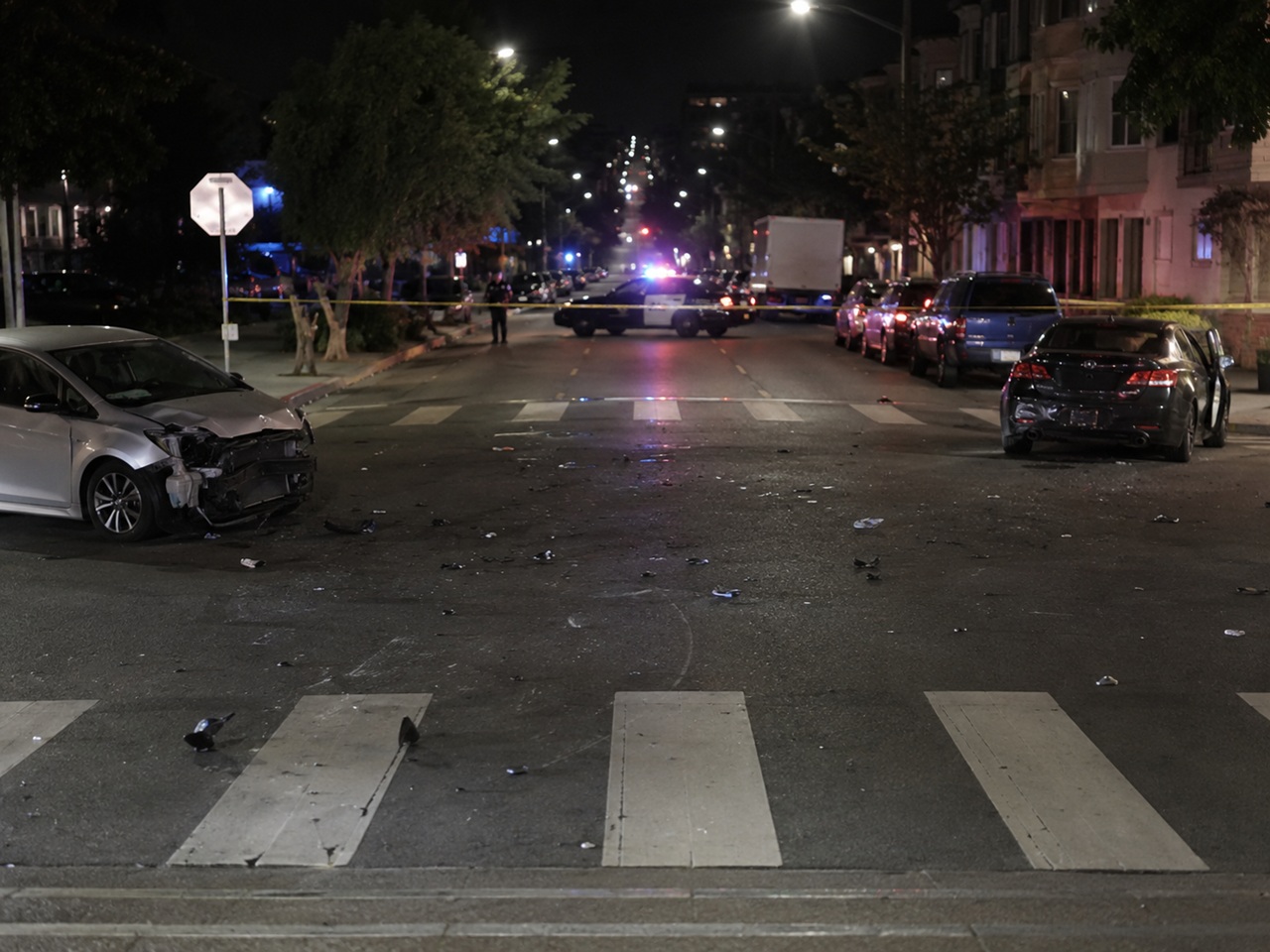 A night scene of a serious hit-and-run crash in a Bay Area residential neighborhood, showing a silver sedan with severe front-end damage and a dark sedan with rear-end damage positioned near a crosswalk covered in debris, with emergency police vehicle lights visible behind a street blockade in the background.