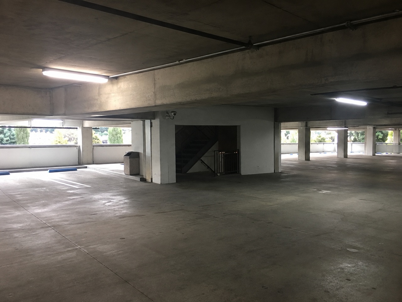 A dimly lit and empty multi-level parking structure in California with a single security camera mounted on a concrete pillar near a dark stairwell entrance, depicting the type of poorly secured commercial property where negligent security claims may arise after an assault, robbery, or other violent incident.